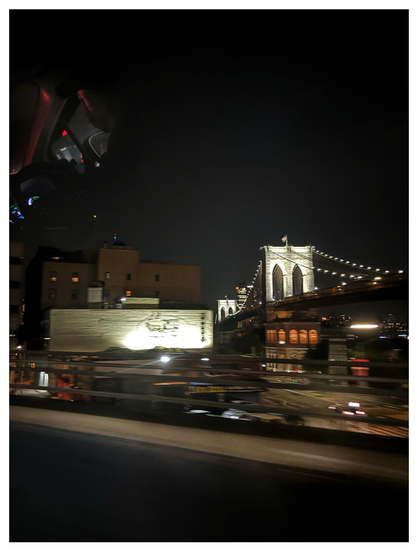 A city scene at night, photographed from behind a car’s glass window. The elevated highway in the foreground rushes past in a blur, overlooking another highway with some vehicles blurred with motion. In the middle distance to the left are some low-rise buildings illuminated by streetlights. Farther off to the right is the Brooklyn Bridge with lights strung along its cables. The sky above is featureless and dark. At upper left is a reflection of the car driver, facing away from camera, and the car’s dashboard lights.