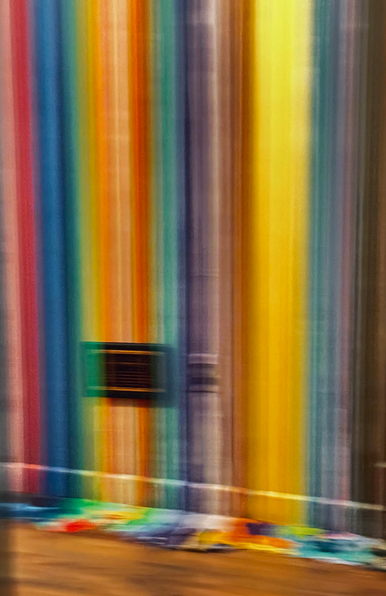 Color photo of a large wall that has been covered in long vertical drips of paint, coming straight down and pooling on the floor. I believe it is the front office of a design firm that is in the building where I had an appointment. As the elevator doors opened I got this blurred shot. It is slightly on an angle and shows blurred vertical stripes of pink red blue orange teal purple brown yellow blue and lavender coming to a blurred mess on the floor. The rest of the floor is plain wood and about a foot up from the bottom of the wall is a black plaque that is unreadable