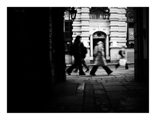 Black and white street photograph using intentional motion blur showing three people walking past ornate Victorian/Georgian stone architecture with classical columns and decorative details, creating dynamic movement against static building features.