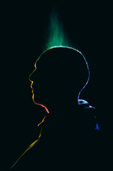 A photograph of a silhouette of a person's (Peter) profile against a black background, illuminated by a colourful light gradient. The gradient starts with green at the top, transitioning through yellow and red to blue at the bottom. The light creates a neon outline around the contours of his head and shoulders, emphasizing his profile