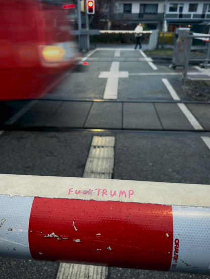 das foto zeigt einen bahnübergang mit einer heruntergelassenen schranke. auf der schranke steht mit pinkem stift "fuck trump" geschrieben. im hintergrund links oben ist eine rote straßenbahn in bewegung zu sehen. die schranke ist rot-weiß gestreift. der bahnübergang ist mit weißen kreuzen und linien markiert. im hintergrund an der anderen schranke ist eine andere person zu sehen.. eine rote ampel leuchtet ebenfalls im hintergrund.