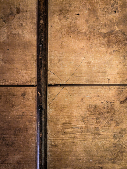 A top-down view of the surface of a weathered, aged wooden chest trunk. The wood has a warm, honey-brown base tone. It features a patina of dark grime, scattered water spots, and numerous fine scratches.

A dark, rounded wooden trim or support beam runs vertically down the left side. It is held in place by visible, dark metal nails or rivets.

A prominent "X" is etched into the wood near the center, appearing almost like a measurement mark or a crude "sign-off" from a past craftsman. 