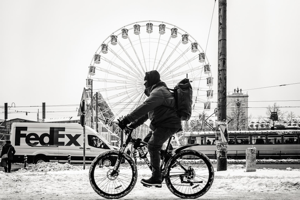 This is a black and white photograph taken from a slightly low angle, capturing a winter street scene. The main subject is a person riding a bicycle, his head centred with a ferris wheel in the background. The cyclist is wearing a hooded jacket and a backpack, obscuring most of their facial features with a dark covering, giving them an anonymous, perhaps hurried appearance. 

Dominating the background is a large Ferris wheel, centrally placed and filling a significant portion of the upper half of the frame. Its intricate spokes and numerous passenger gondolas are clearly visible against a bright, overcast sky. 