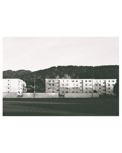 Black and white photograph. In the front a wide lawn, supposedly a football field, that ends at the back of white concrete walls with two floodlights. Behind the walls a small block of flat estates. In the background wooden hills and a bright sky with a slight redish shine. The sunlight comes from the right and is carving some shadow/light play onto the lawn and the flat surfaces.