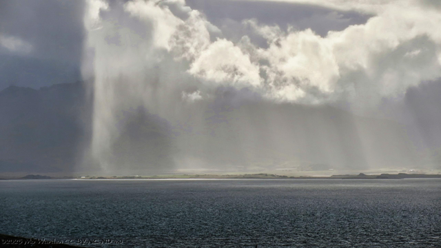 A colour photo of a seascape with the curves of a hazy mountain range in the distance. The outline of the seashore has been picked out by sunlight striking through a rain shower. A few buildings are just visible. Low cloud is being highlighted by the sunbeams, as well as the soft random fall of rain. It's very beautiful.
