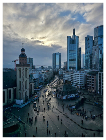 An elevated, high-angle view of a bustling city square and street on a cloudy day. In the foreground, a dark, wet pedestrian plaza is scattered with people walking. To the left, a tall, historic church tower with a clock and a domed top stands prominently. In the middle ground, a narrow street lined with multi-story buildings and parked cars curves toward the background. A small, traditional building with a dark roof sits in the center of the plaza. The background is dominated by a skyline of modern glass skyscrapers, including one notably tall building with a spire, set against a dramatic, overcast sky where the sun is faintly breaking through the clouds.