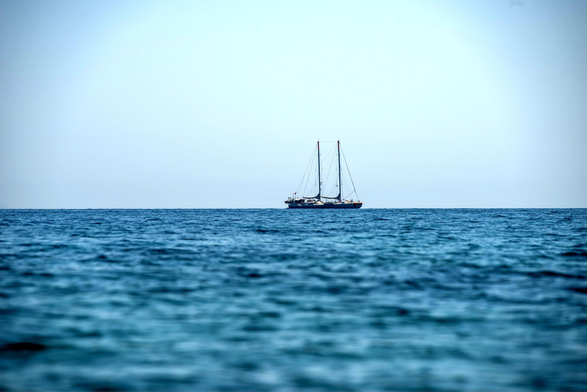 Das Bild zeigt eine weite Ansicht des Meeres und des Himmels, die durch den Horizont klar in zwei Hälften geteilt ist. Im Vordergrund dominiert das tiefblaue, leicht wellige Wasser, das die untere Bildhälfte ausfüllt. Weiter hinten, genau auf der Horizontlinie in der Mitte des Bildes, ist ein zweimastiges Segelboot zu sehen. Das Boot wirkt klein in der Weite der Szenerie. Der obere Teil des Bildes zeigt einen sehr hellen, fast weißen bis hellblauen Himmel, der nahtlos in den blauen Ozean übergeht. Die Beleuchtung ist hell, was auf einen sonnigen Tag hinweist. Die Szene strahlt Ruhe und Weite aus.
