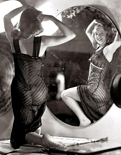 Marilyn Monroe portant un déshabillé transparent posant devant de trois-quarts dos devant un grand miroir, prétendant parfaire sa coiffure et offrant l’image de son corps de starlette aux voyeurs