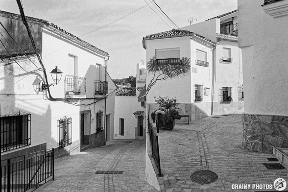 A charming narrow street in Benadalid, featuring traditional balconies and a cobblestone path. The scene is serene, with a few potted plants and a vintage street lamp adding to the tranquil atmosphere.