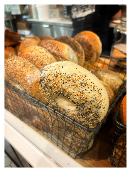 “Everything” bagels stacked upright in rows, in a wire basket on a bakery counter. The bagels are plump and a toasted brown color, coated with garlic, onion, and poppy and sesame seeds. Taken at Mazzola Bakery in Brooklyn.