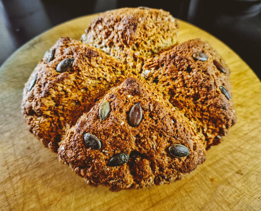 A loaf of Irish soda bread sits on a wooden cutting board. The loaf is golden brown with a rough, uneven texture, and has been scored with a cross on it. A generous amount of pumpkin seeds are on the top. The cutting board is light brown and has visible grain. The background is dark and out of focus.