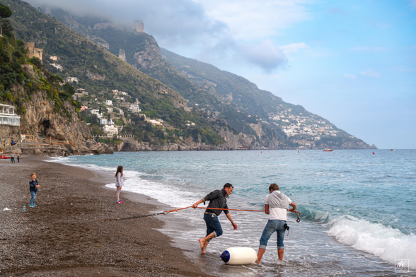 Color photo of two people hauling a rope on a pebbly beach with two young people on the beach beyond and cloud-topped mountains with many houses on them rising from the sea in the background.