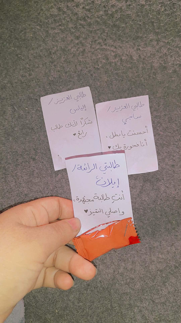 Hand holding a small orange candy wrapper, underneath three handwritten notes in Arabic praising students, on a gray textured surface.