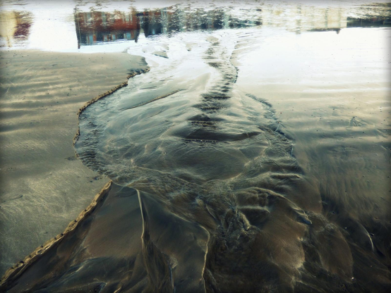 A colour photograph showing a water outflow on wet sand with reflected buildings at the top of the frame and the outflow passing vertically through the centre of the frame.