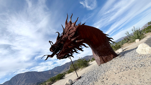 A metal sculpture of the head of a dragon/snake at the Galleta Meadows sculpture exhibit in Borrego Springs California. The sculpture is rust coloured and at least 6m/20ft high. The soil around the dragon is bare with a some green vegetation in the background. At the horizon you can see some hills. Horizon of the photo is angled; low on the left, higher on the right. Sky is blue with some clouds.