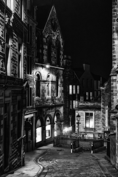 Eine enge, gepflasterte Gasse in Edinburghs Upper Bow schlängelt sich durch historische Steingebäude bei Nacht. Die Szene wird von Strassenlaternen und beleuchteten Fenstern erhellt, die starke Kontraste und tiefe Schatten auf die rauen Fassaden werfen. Die Schwarz-Weiss-Darstellung betont die architektonischen Details und die atmosphärische Tiefe der urbanen Landschaft.