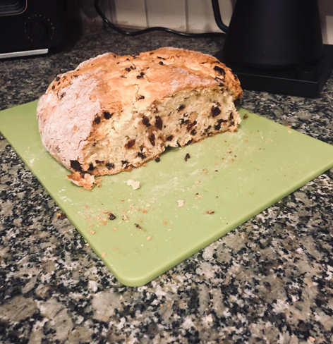 My nice round loaf of Irish soda bread with currants (raisins), a quarter of it has been cut off so you can see all of the currants inside. It is sitting on a lime green cutting board which is on a green and black speckled granite type countertop. In the back can be seen bits of my water kettle and toaster
