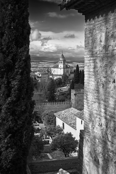 view from a secluded spot at the Alhambra. At left is a tall cedar and at right is one of the ancient structures. In the middle is an opening that provides a vista over the complex, to the city of Granada, and to the countryside beyond. Low clouds cover the valley but there is blue sky above. 