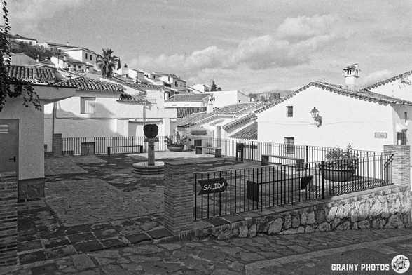 A quiet village square featuring traditional whitewashed buildings with tiled roofs, a stone fountain at the center, and a sign indicating "SALIDA". The scene captures the tranquil atmosphere of a historic neighborhood, set against a cloudy sky.