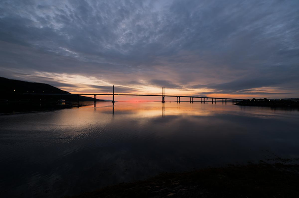 Eine Brücke führt über eine Meerenge. Es ist kurz vor Sonnenaufgang. Genau an der Stelle des Sonnenaufgangs gibt es keine Wolken. Der Rest ist komplett mit Wolken bedeckt. Im Vordergrund ist Wasser.