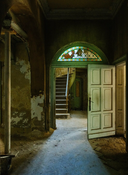 An abandoned interior space featuring a staircase, two doorways, and stained glass above the entrance. The walls show signs of decay, with peeling paint and dust on the floor.