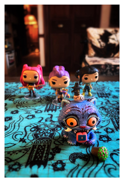 Plastic Funko figurines of characters from the animated Netflix film “K-Pop Demon Hunters.” In the foreground to the right is Derpy, a purple mythological Korean tiger with wild-looking orange eyes and a drooping tongue with an envelope stuck to it. In front of him is a flower pot tipped over on its side, a reference to a scene from the film. Perched on his head is Sussie, a six-eyed magpie wearing a small black hat. Lined up behind Derpy are three female figurines with differently colored hair: Mira, Rumi, and Zoey, the three members of Huntrix, posing with their weapons. The figures are arranged on a blue-dyed fabric with black batik patterns. In the background is a blurred living room scene: a chair with a cushion, a closet door, a lamp down a hallway.