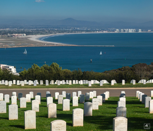 Color photo looking down a slope filled with the rows of white tombstones of Fort Rosecrans National Cemetery, with San Diego harbor and Coronado Island below and beyond. On the far horizon are the hills and mountains of northern Baja Mexico. 