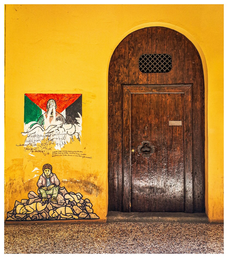 A vibrant yellow wall featuring poignant street art depicting human suffering and symbols of the Palestinian struggle stands beside an old, dark wooden arched door. 
The first piece of street art is of a small child sitting on top of rubble with outlines of bodies under them. The second piece above is of a Palestinian flag with writing in English and in Arabic on/under it.