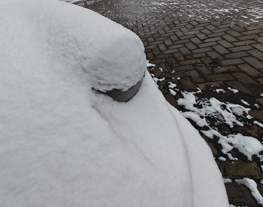 A photo of the front of a  parked car almost completely covered in snow, with a single round headlight partially visible. A brick-paved street can be seen in the background.