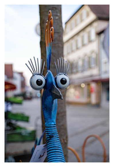 Foto im Hochformat. Nahaufnahme eine Figur aus Metall. Es soll wohl ein Pfau sein, im Bild ist der blaue gebogene has, oben der Kopf mit Schnabel. Beide Augen sind seitlich am Kopf und kugelrund aufgerissen. Darüber lange Wimpern. Auf dem Kopf ist ein blau-orangener Hahnenkamm. Am Hals hängt ein Preisschild. Im Hintergrund ist unscharf eine Fußgängerzone.
