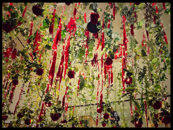 A color photo that was taken straight up towards the ceiling in a café. To decorate, they have hung very long strings of fake greenery, ending with a fake upside down rose. There are also red ribbon and fairy lights woven into some of the strings. There's quite a lot of them, quite close together so it's a very crowded photo. Behind the strings of greenery can be seen a few black poles and a table from an upper floor in the café. A black border has been added to the photo that mimics the black bars a bit