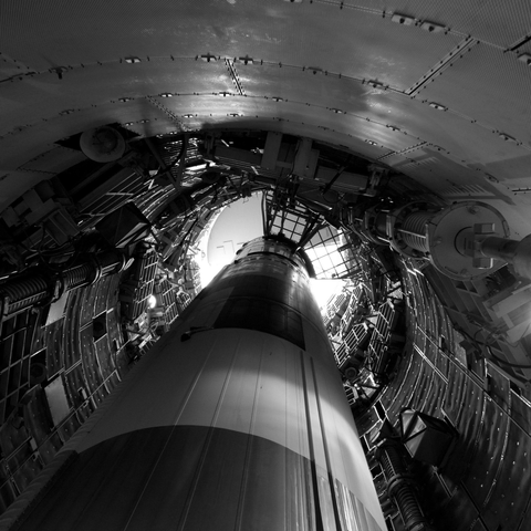 Looking up from the bottom of a missile pointed up inside a cramped underground silo, which is lined with retractable platforms and various apparatus.