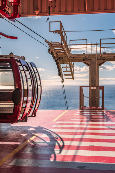 Das Bild zeigt eine Gondelstation am Meer an einem sonnigen Tag.

Im Vordergrund steht eine rote Gondel mit abgerundeten Glasfenstern auf einer rot-weiß gestreiften Plattform. Die Plattform hat gelbe Markierungen und ist durch den Schatten einer nicht sichtbaren Struktur verdeckt. Hinter der Gondel erstreckt sich ein strahlend blaues Meer bis zum Horizont unter einem hellblauen Himmel mit einigen vereinzelten Wolken. Rechts von der Gondel befindet sich ein metallischer Gondelträger mit Treppen, der in den Himmel ragt und die Gondelkabel hält. Die Plattform, auf der die Gondel steht, ist mit rot-weißen Streifen und einigen gelben Markierungen versehen.