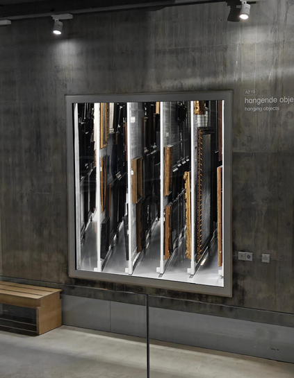 A photo looking at an inside windown in a museum. Behind the window one can see multiple framed artworks stored vertically on sliding racks. The style of the building is bare concrete walls and spot lighting.