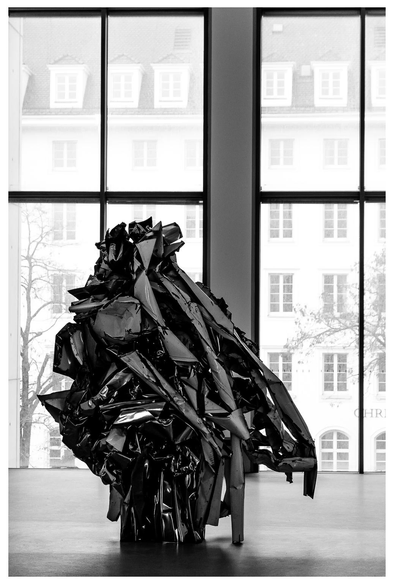 Eine sehr amorphe Skulptur - wenn man es so nennen darf - aus Schrotteilen steht vor einer großen Fensterfront durch die man ein Haus mit - überraschung - Fenstern sieht.