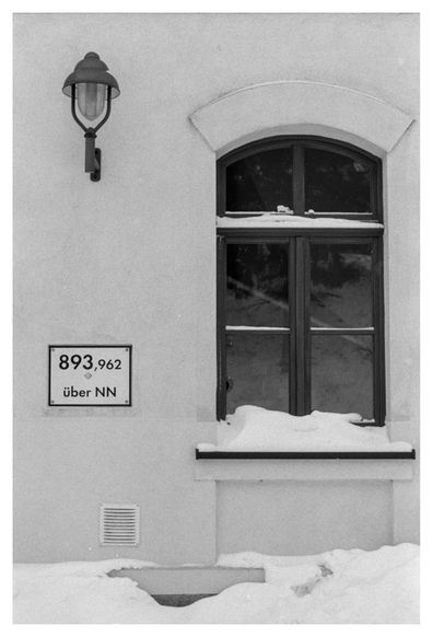 Black and white photograph in portrait orientation. A window at a train station with a sign indicating the height: almost 894 m. There is snow on the ground and the window sill.