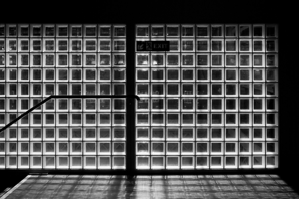 das bild ist in schwarzweiß gehalten und zeigt eine wand bzw. ein sehr, sehr großes fenster aus glasbausteinen. in der mitte, etwas nach oben versetzt, befindet sich ein ausgangsschild mit der aufschrift "exit" und einem pfeil, der nach links unten zeigt. links ist ein stück geländer einer treppe zu sehen, der nach unten führt. der boden vor der wand reflektiert das licht, das durch die glasbausteine scheint, und erzeugt ein interessantes schattenspiel. das bild hat eine hohe kontrastwirkung.