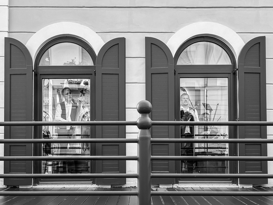 Zwei hohe Fenster, Erdgeschoss und bodennah, mit geöffneten Fensterläden. In der Spiegelung der Scheiben erkennt man Musiker mit Instrumenten, die mit dem Rücken zum Raum stehen.
Schwarzweiß.