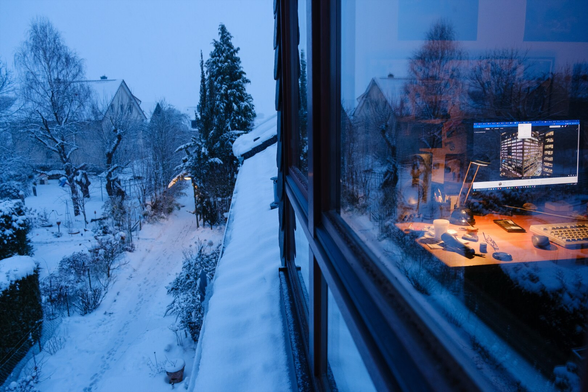 Eine verschneite Außenszene wird durch ein Fenster eingefangen und spiegelt einen warm beleuchteten Innenarbeitsraum wider. Der verschneite Garten verfügt über Bäume, Sträucher und einen Weg, während sich im Inneren ein Schreibtisch mit verschiedenen Gegenständen und eine Lampe befindet, die sanft gegen den sich verdunkelnden Himmel leuchtet.