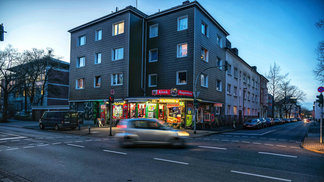 Das Bild zeigt eine Straßenecke in einer Stadt während der Abenddämmerung. Im Zentrum des Bildes steht ein vierstöckiges Gebäude mit einer dunkelgrauen Fassade. Im Erdgeschoss dieses Gebäudes befindet sich ein beleuchteter Kiosk und eine Baguetteria, deren rote und gelbe Schilder sichtbar sind. Mehrere Fenster in den oberen Stockwerken sind ebenfalls beleuchtet. Im Vordergrund, auf der Straße, fährt ein silbernes Auto von links nach rechts durch das Bild; es ist aufgrund der Bewegungsunschärfe verschwommen dargestellt. Links neben der Fahrbahn ist ein schwarzer Van geparkt. Die Straßen sind durch weiße Linien markiert. Rechts erstreckt sich eine weitere Straße mit parkenden Autos am Straßenrand und mehreren Laternen, die die Szene erhellen. Im Hintergrund links ist ein weiteres, höheres Gebäude zu sehen. Der Himmel ist in Blautönen gehalten, und es sind einige kahle Bäume an den Rändern der Straßen sichtbar.