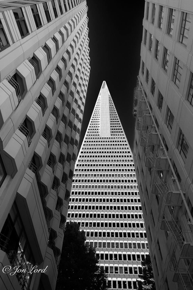 This is a black and white cityscape photo of a tall, triangular shaped high-rise, that, with the perspective of distant converging lines gives the impression that the top of the building disappears into infinity (space). Transamerica Building (2018).

On either side of the photo are two rectangular high-rise buildings, that with optical distortion, give the impression that the two buildings lean inwards slightly towards the centre. Both buildings are light or pale grey. The left is the more modern with multiple 'bay' shaped balconies, so perhaps residential or a hotel. The right older block has numerous evenly spaced rectangular windows with an external fire escape and pipework. In the centre of the image is our tall, triangular, high-rise office block. The camera is angled upwards towards the upper floors. The building is brilliant white with tall rectangular windows. It tapers evenly and sharply with a pointed top. With the perspective of 'converging lines' the tip of the tower seems to go on forever and finish in space, if at all. The sky around the summits of the buildings is clear and cloudless, rendered jet black and devoid of any detail. 

The Building is the 48 story, 260m tall, Transamerica Pyramid in San Francisco's Financial District.

