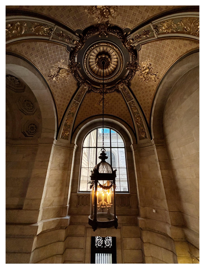 A view from the cathedral-like interior of the entrance hall of the New York Public Library’s main research branch at 42nd Street. Massive marble walls are flanked by soaring arches, from which wide bands with bas-relief elements rise and converge on a series of concentric rings at the top of a heavily ornamented domed ceiling. Hanging by a long chain from the center of the rings is a cylindrical lantern in a glass and metal casing with a domed top, containing several glowing electric candles. Behind the lantern, and framing it, is an arched window with a view of the outside, where we can glimpse another wing of the library with its own matching window.