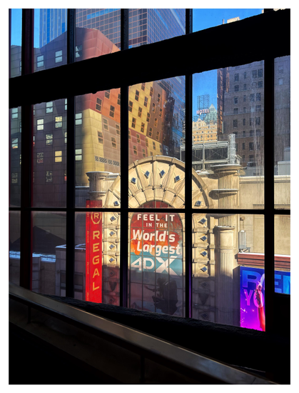 A view of city buildings, broken up into rectangles by a grid of tall glass windows. The photo is taken from a darkened interior, with a light-colored guardrail for an ascending ramp in the foreground. The main building in view is a multistory edifice with an Art Deco facade, featuring a massive arched frame lined with little diamond-shaped glass panels. This frame surrounds a large central arched window covered with a giant sign that reads “FEEL IT IN THE WORLD’S LARGEST 4DX.” The window is flanked by faux Greek columns and some vividly colored digital displays, including a neon-red vertical sign that says “REGAL.” Beside the Regal multiplex is an oddly shaped trapezoidal building, with a checkerboard pattern of windows set in a facade with differently colored rectangular sections.