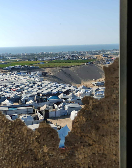 Overhead view through a broken window of a refugee camp comprised of numerous white tents, with a coastal city and ocean in the background.