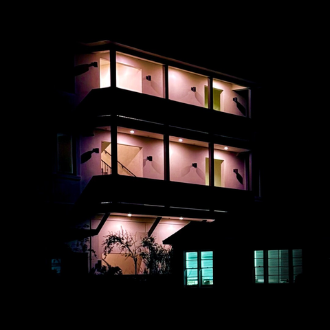 Nighttime shot of a large apartment building cast in a pinkish hue with a much smaller home in the foreground with two windows illuminated in light blue
