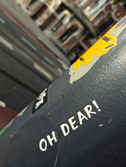 A close-up photo of a worn black pole with peeling stickers and the white text “OH DEAR!”, with a city street at an angle in the background.