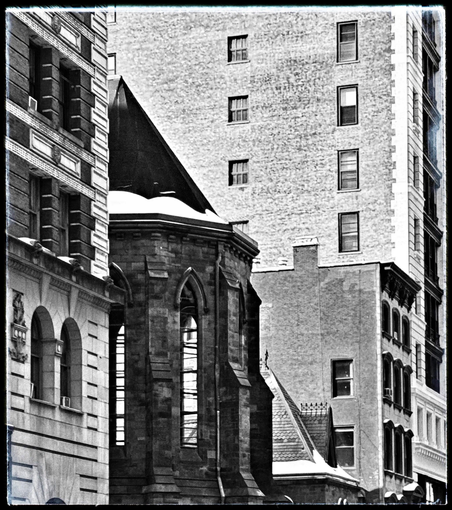 A black and white photo taken at an angle down 26th St. showing the side of a church in between two other buildings. It is the Serbian Orthodox Church on 26th and Broadway. In 2016 it had a major fire, no one was hurt, but the inside was gutted and has not been renovated. The church has a black roof that is going up into a point with snow sitting on the balustrade. Below that it is curved stone work with long thin windows that arch at the top. The windows are empty so you can see straight through to the building behind it. On its left is an older stonework building with two rounded windows and on the right is a taller plain brick building with a few windows on the side