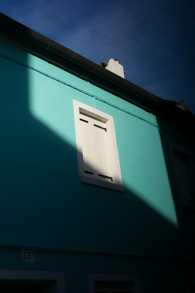 Photo looking up at a mint green house under a lightly dusted blue sky. Strong sunlight illuminates the top half, but dark shadows from other buildings obscures the bottom. In the middle is a single small bright white window, shutters tightly closed.