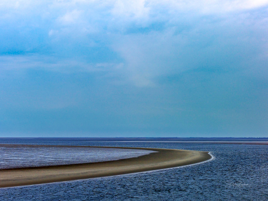 A serene view of a curved sandy beach extending into calm blue waters under a cloudy sky. The landscape features a subtle gradient from land to sea, highlighting the tranquility of the coastal scene.