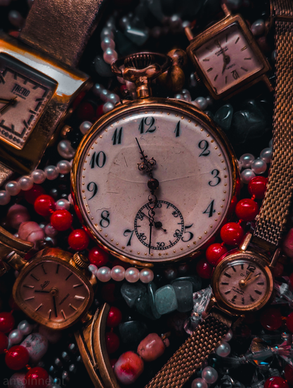 A rich, moody, close-up of several vintage timepieces nestled among various necklaces made out of beads and stones. The "still life" evokes a sense of nostalgia, the passage of time, or a collection of forgotten treasures.

Occupying the center is a large, gold-toned pocket watch with a white, slightly weathered face. It features elegant black Arabic numerals and intricate, ornate hands. There is a smaller sub-dial at the bottom for seconds.

Surrounding the pocket watch are smaller vintage wristwatches. They vary in shape, square, oval, and round with gold-toned casings and weathered faces. The watches are resting on a bed of strands of white pearls, vibrant red spherical beads, sea-glass green stones and clear crystalline fragments. 

The deep reds and golds pop against a very dark, shadowed background.